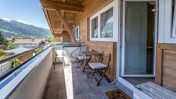 Un balcone in legno con un tavolo e sedie, una porta scorrevole in vetro e vista sulle montagne.