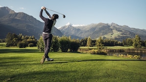 Uma pessoa jogando golfe num campo bem cuidado, com um lago e montanhas ao fundo.