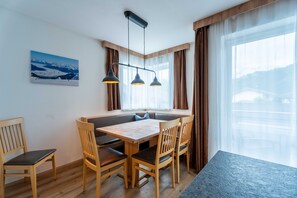 Un comedor con mesa y sillas de madera, una lámpara colgante, una ventana con cortinas y un cuadro con paisaje de montaña en la pared.