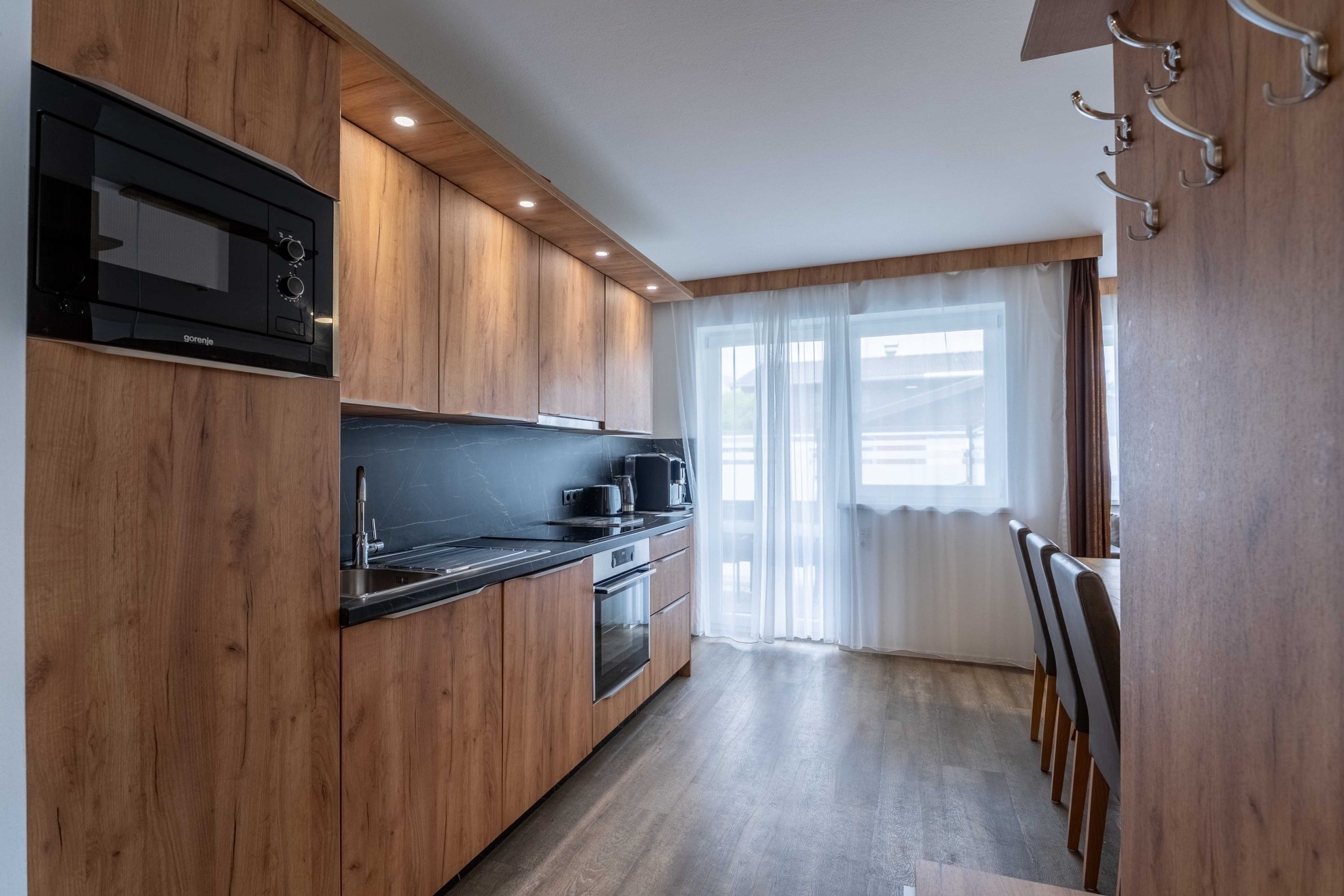 A modern kitchen with wooden cabinets, a black countertop, a built-in microwave, and a window with sheer curtains.