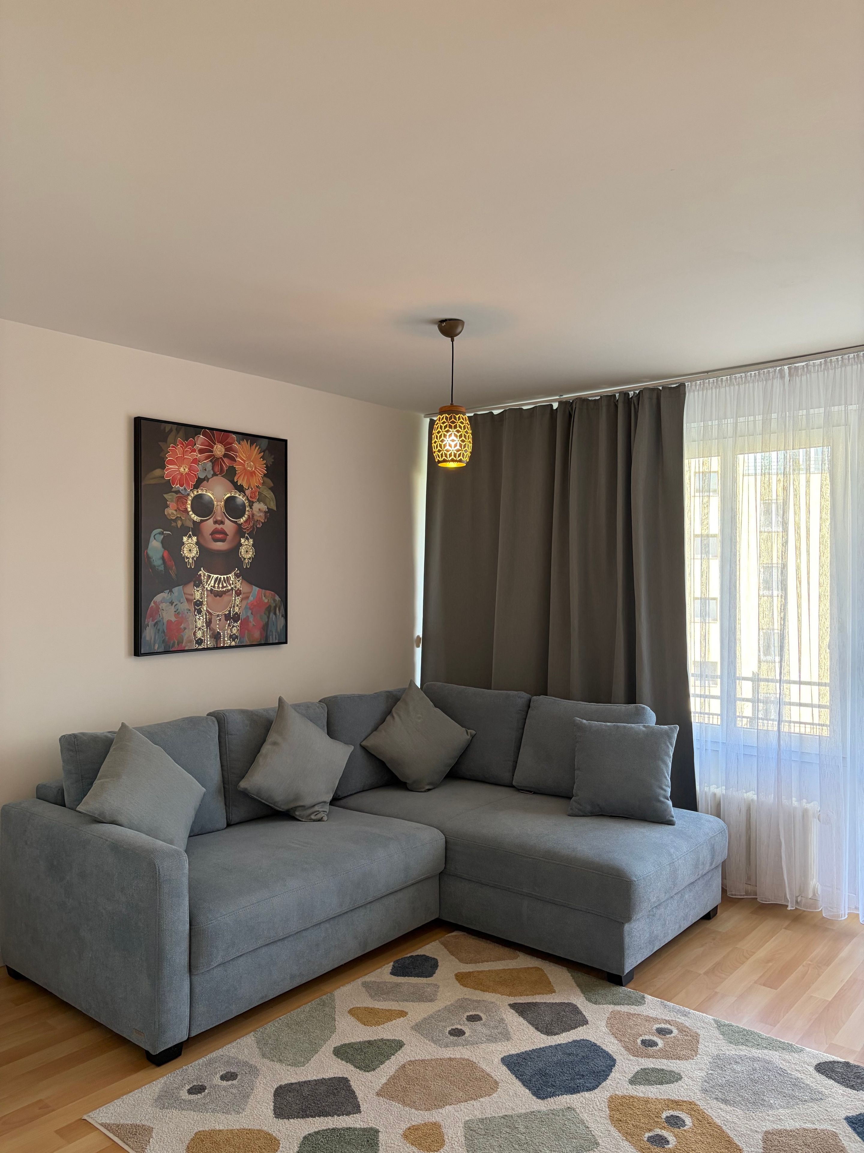 Een moderne woonkamer met een grijze hoekbank, een kamerkleed met dessin, een hanglamp en een schilderij aan de muur.