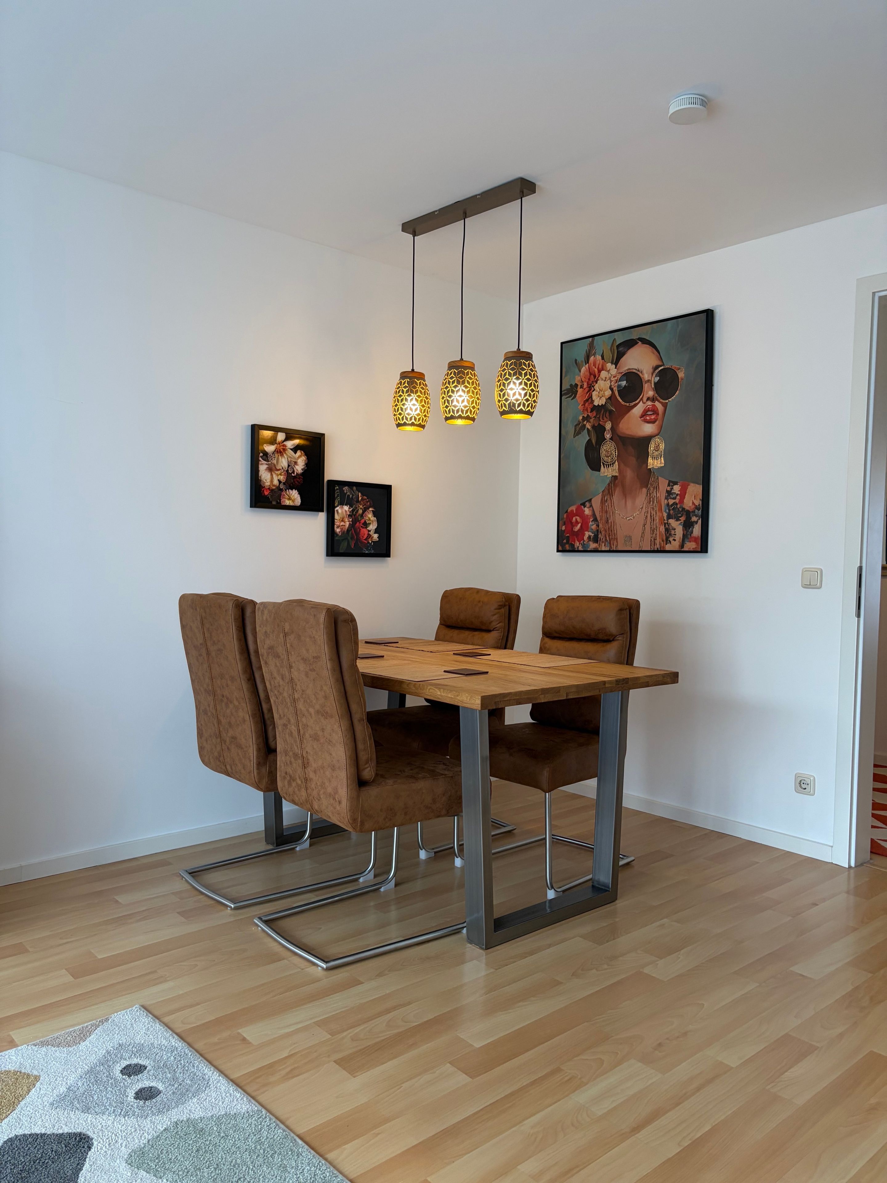 A dining area with a wooden table and brown chairs, three hanging pendant lights, and two framed floral paintings on the wall.
