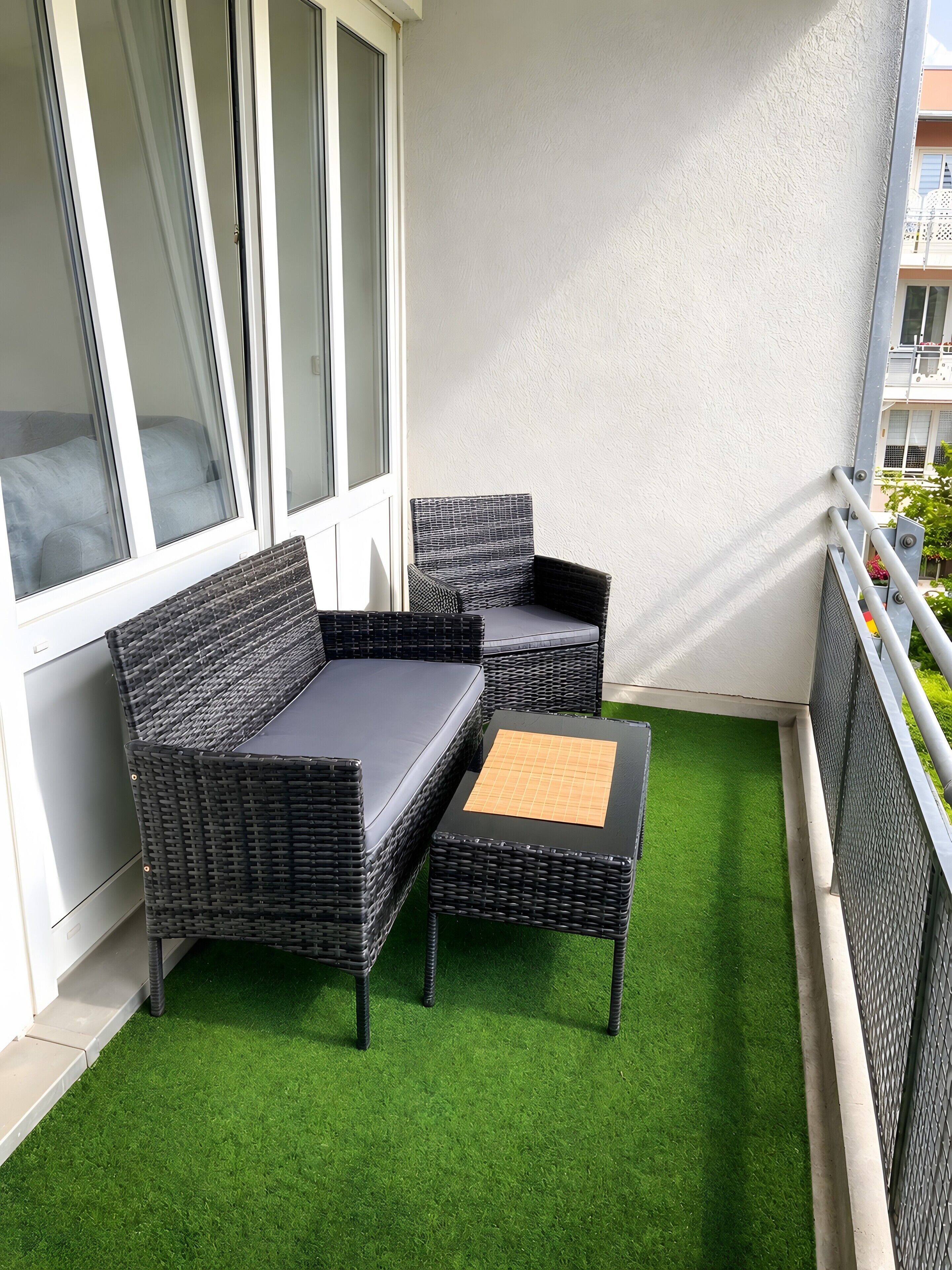 A balcony with a black wicker sofa and a matching ottoman, a small table, and a green artificial grass floor.