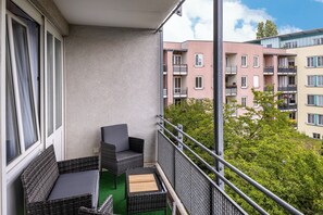 Een balkon met rieten meubilair, een tafeltje en uitzicht op omliggende gebouwen en groen.