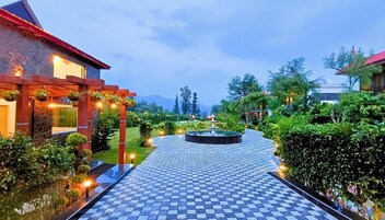 Entrada de hotel com um caminho quadriculado que leva a uma fonte circular, cercada por vegetação exuberante e iluminada por iluminação ambiente.
