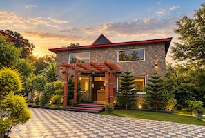Una casa de dos pisos con fachada de piedra, techo rojo y una pérgola de madera con macetas colgantes.