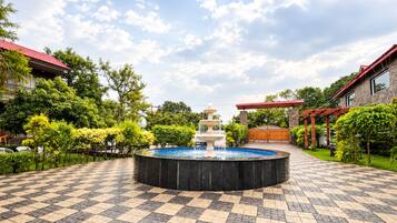 A courtyard with a central fountain, surrounded by a checkered tile floor, greenery, and a building with a red roof.