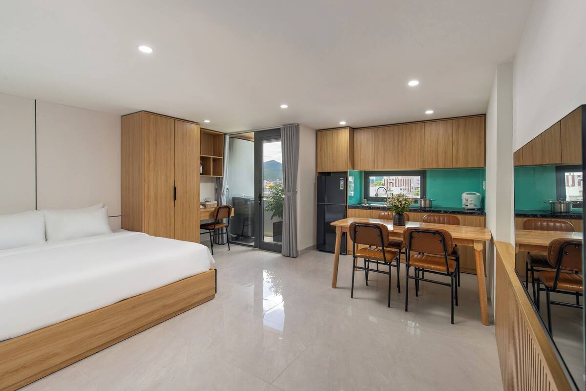 Een moderne hotelkamer met een bed, een zithoek met stoelen, een keuken met een koelkast, en uitzicht door een glazen deur.