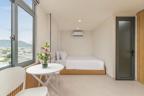 A modern bedroom with a bed, a small table with a vase, a chair, and a view of the city through large windows. - Ngoc Thuong Hotel & Apartment (An Hai)