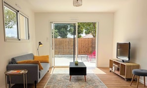 Una sala de estar con un sofá, una mesita, un televisor y un ventanal corredizo que da a un patio.