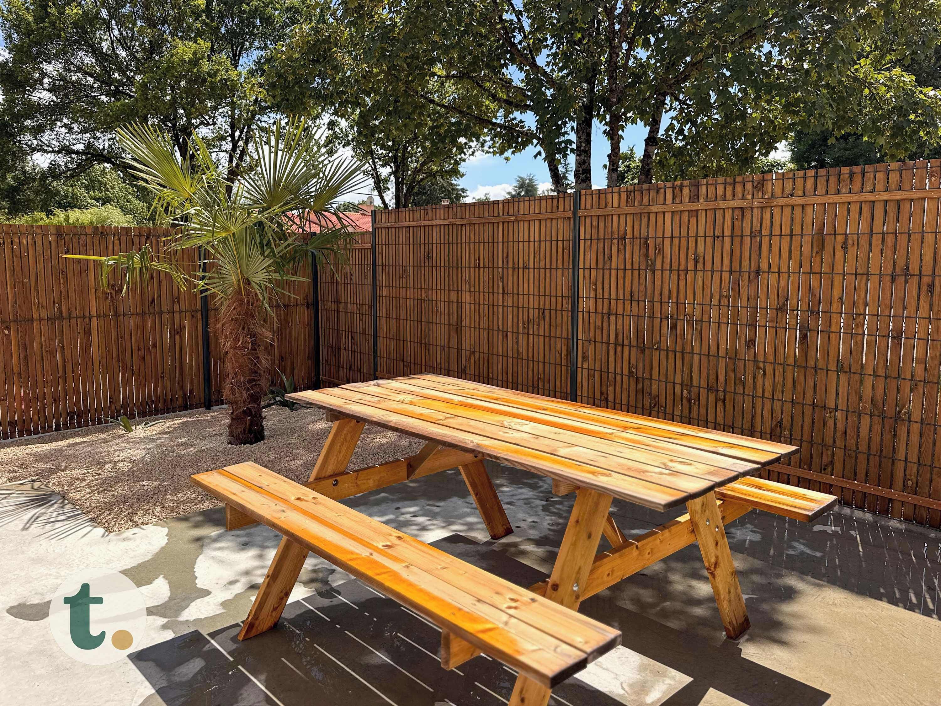 A wooden picnic table with benches in a backyard with a wooden fence and trees.