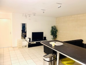 A modern living room with a flat-screen TV, a dining table, a sofa, a potted plant, and a trash bin. - Quiet 3-room apartment in Gradignan, near Bordeaux (Gradignan)
