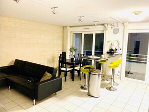 A modern living room with a black sofa, dining area with chairs, a kitchen counter, and a trash can. - Quiet 3-room apartment in Gradignan, near Bordeaux (Gradignan)