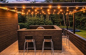 Ein Außenbereich mit Grillstation, zwei Barhocker und Lichterketten.