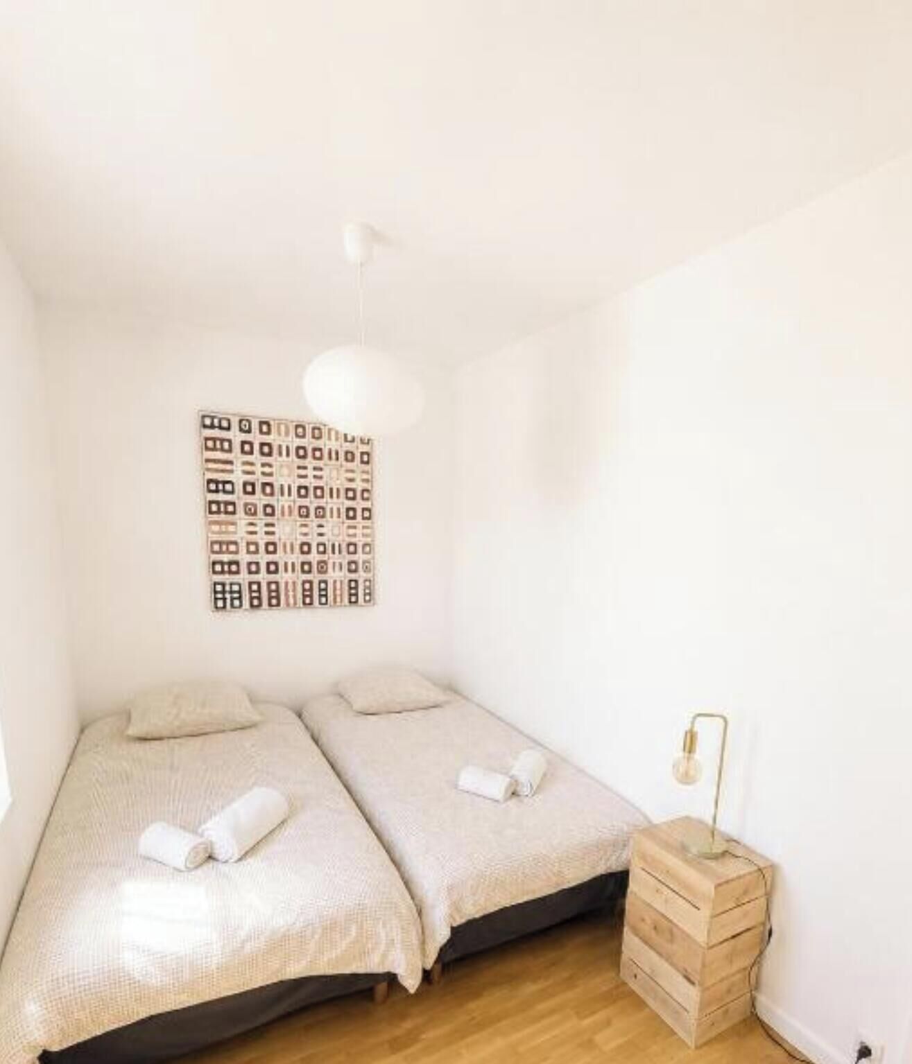 Une petite chambre propre, avec deux lits, une œuvre d’art murale, une lampe de chevet et une table de chevet en bois.