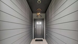 A narrow hallway with grey siding walls, a door with a window, and a hanging light fixture. - Texas Crew Retreat:4BR, Huge Parking & TVs (Cove)