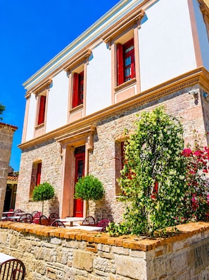 A two-story building with stone walls, red shutters, and a small garden area with potted plants and flowers. - Konak 1882 (Ayvalik)