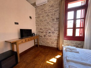 A room with a wooden desk, a television, a stone wall, a window with red shutters, and a bed with white linens. - Konak 1882 (Ayvalik)