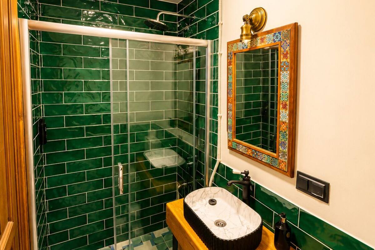 A bathroom with green-tiled walls, a glass-enclosed shower, a wooden vanity with a sink, and a mirror with a decorative frame.