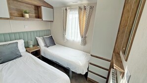 Un dormitorio pequeño y acogedor con dos camas, una mesita de noche con lámpara, una ventana con cortinas y un cabecero de madera.