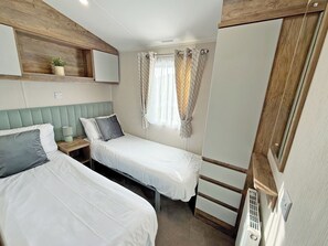 Ein kleines, gemütliches Schlafzimmer mit zwei Betten, einem Nachttisch mit Lampe, einem Fenster mit Vorhängen und einem hölzernen Kopfteil.
