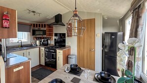 A compact kitchen with modern appliances, a dining area with black tableware, and a potted orchid by the window. - Sunnymede B8 - UKC9571 (Ingoldmells)