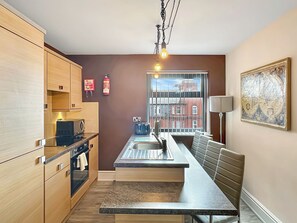 A modern kitchen with a built-in oven, a sink, and a dining area with a bench. There is a fire extinguisher on the wall and a window with a view of buildings outside. - BK302 Billow - UKC9592 (Blackpool)