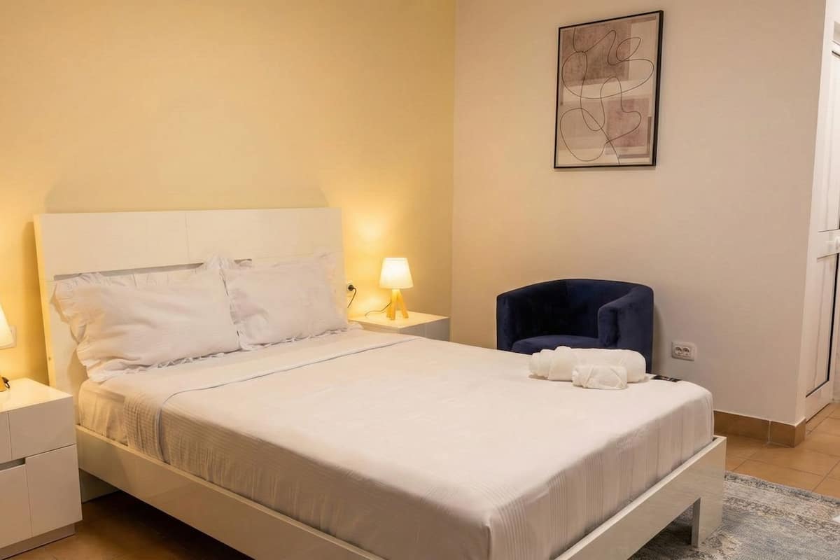 A neatly made bed with white linens, two bedside lamps, a blue chair, and a framed abstract artwork on the wall.