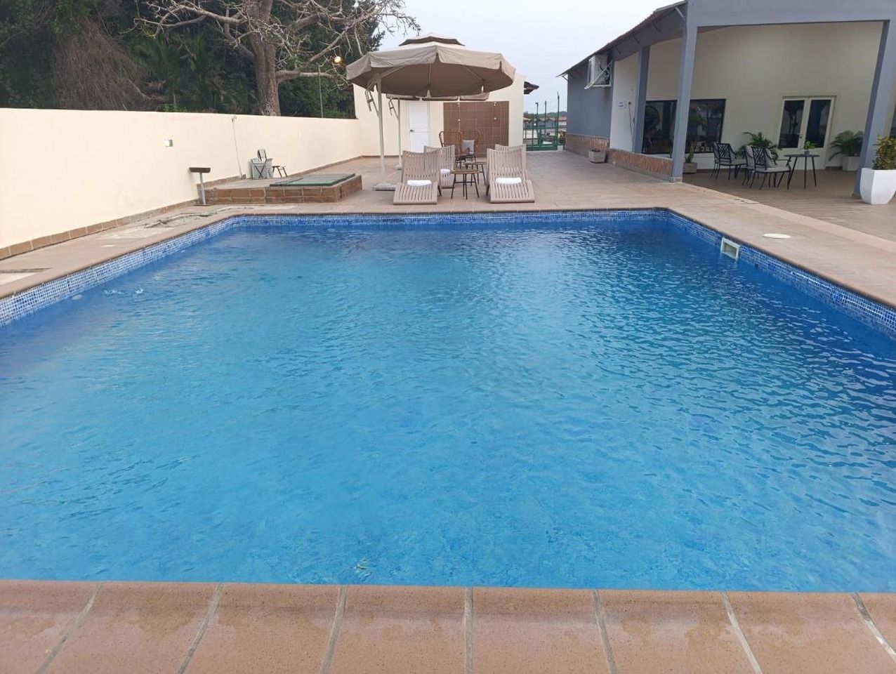 Una piscina rectangular con borde de azulejos, rodeada por una zona de patio con mobiliario exterior y un área de estar cubierta.