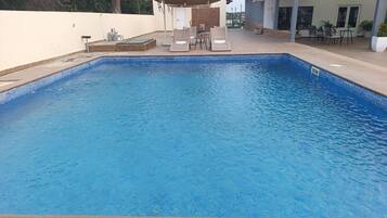 Una piscina rectangular con borde de azulejos, rodeada por una zona de patio con mobiliario exterior y un área de estar cubierta.