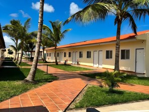 A row of single-story buildings with a red-tiled roof, surrounded by palm trees and a clear blue sky.