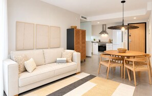 Ein modernes Wohnzimmer mit einer Couch, einem Esstisch mit Stühlen, einer Küche im Hintergrund und einem großen Fenster.