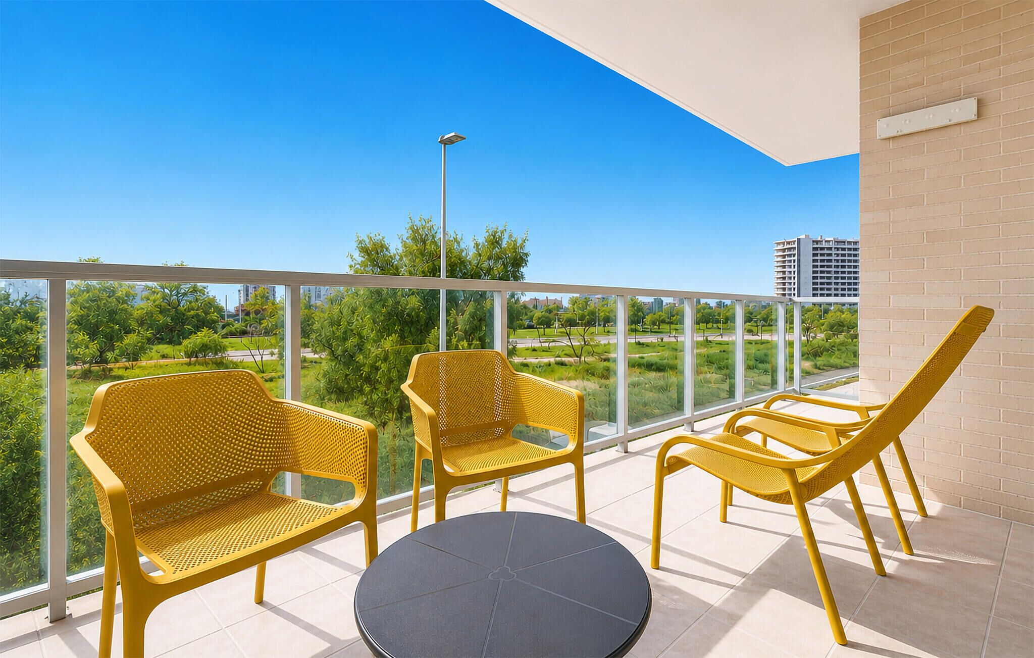 Een balkon met gele stoelen en een ronde tafel, met uitzicht op groen en gebouwen.