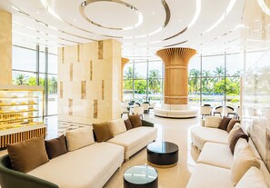 Um hall de hotel moderno com uma área de estar central, uma mesa com sobremesas e grandes janelas com vista para palmeiras.