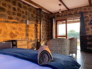 A room with stone walls, a bed with a rolled-up blanket, a wicker chair, and a piano.