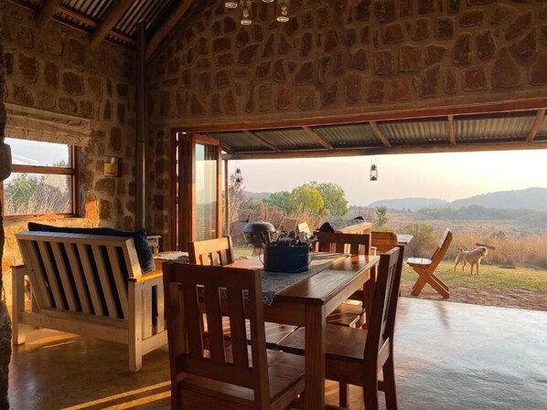 A stone-walled room with a wooden dining table and chairs, a bench, and a view of a landscape with hills and a dog.