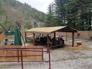 Un espace extérieur aménagé avec des tables et des chaises, abrité par un toit de chaume, et entouré d’une clôture en bois et d’arbres.