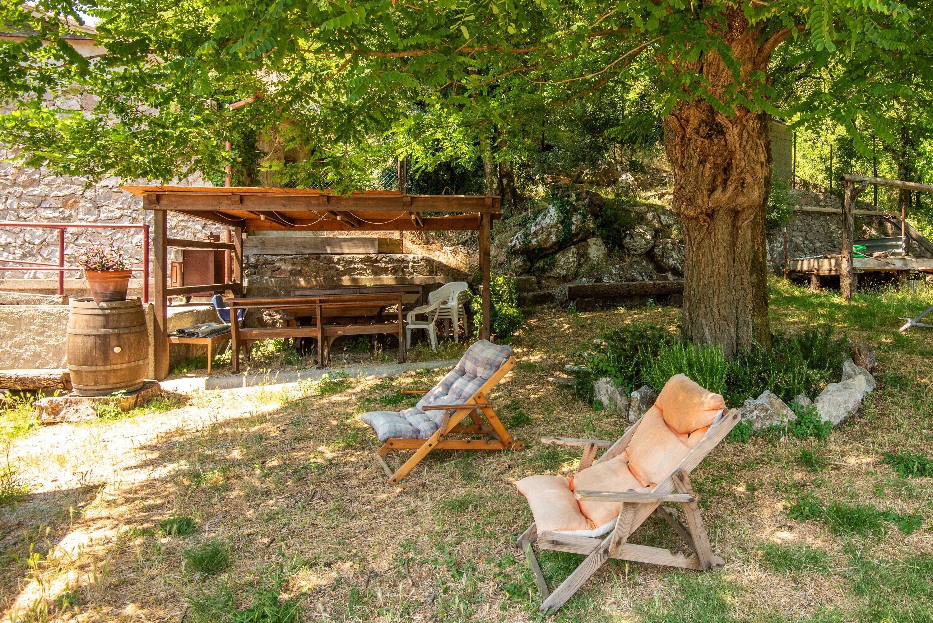 A rustic outdoor seating area with wooden furniture, a pergola, and a large tree providing shade.
