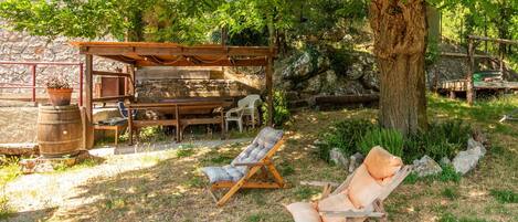 A rustic outdoor seating area with wooden furniture, a pergola, and a large tree providing shade.