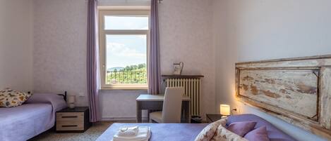 A bedroom with two beds, a desk, a radiator, and a window with curtains.