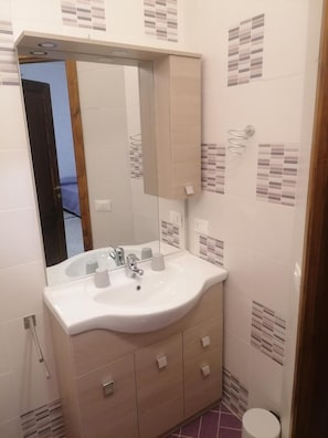 Een badkamer met een wit wastafelmeubel, spiegel en kastje, betegelde wanden en een zichtbaar deurkozijn.