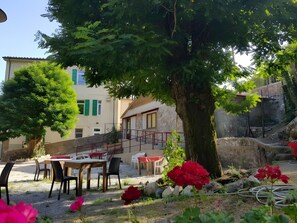 An outdoor seating area with tables and chairs, a tree, and a building with a stone facade in the background.