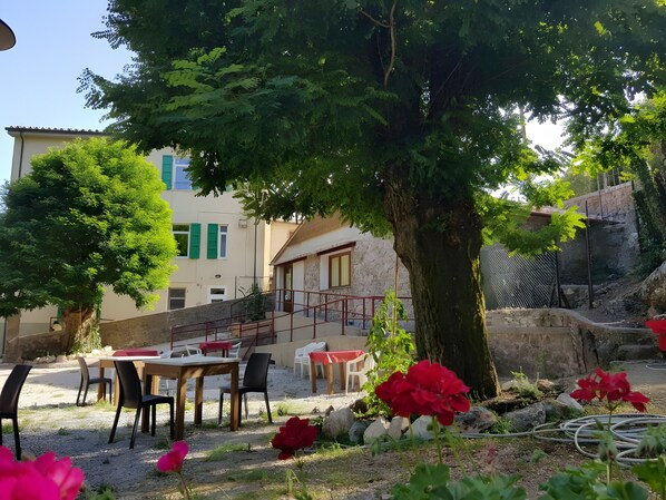 Ein Sitzbereich im Freien mit Tischen und Stühlen, ein Baum und im Hintergrund ein Gebäude mit einer Steinaußenwand.