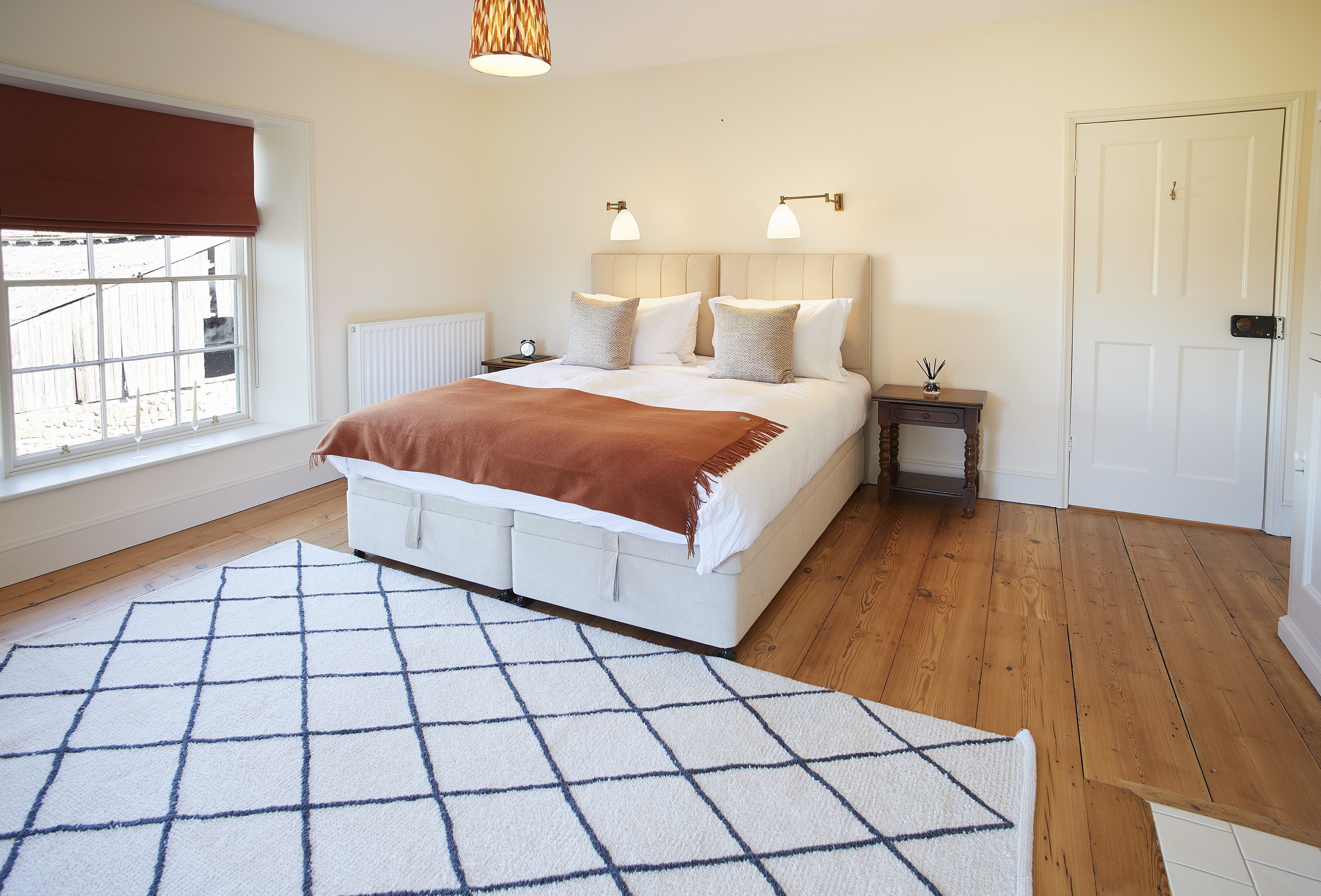 A bedroom with a large bed, a bedside table, a nightstand, a window with a blind, and a door.