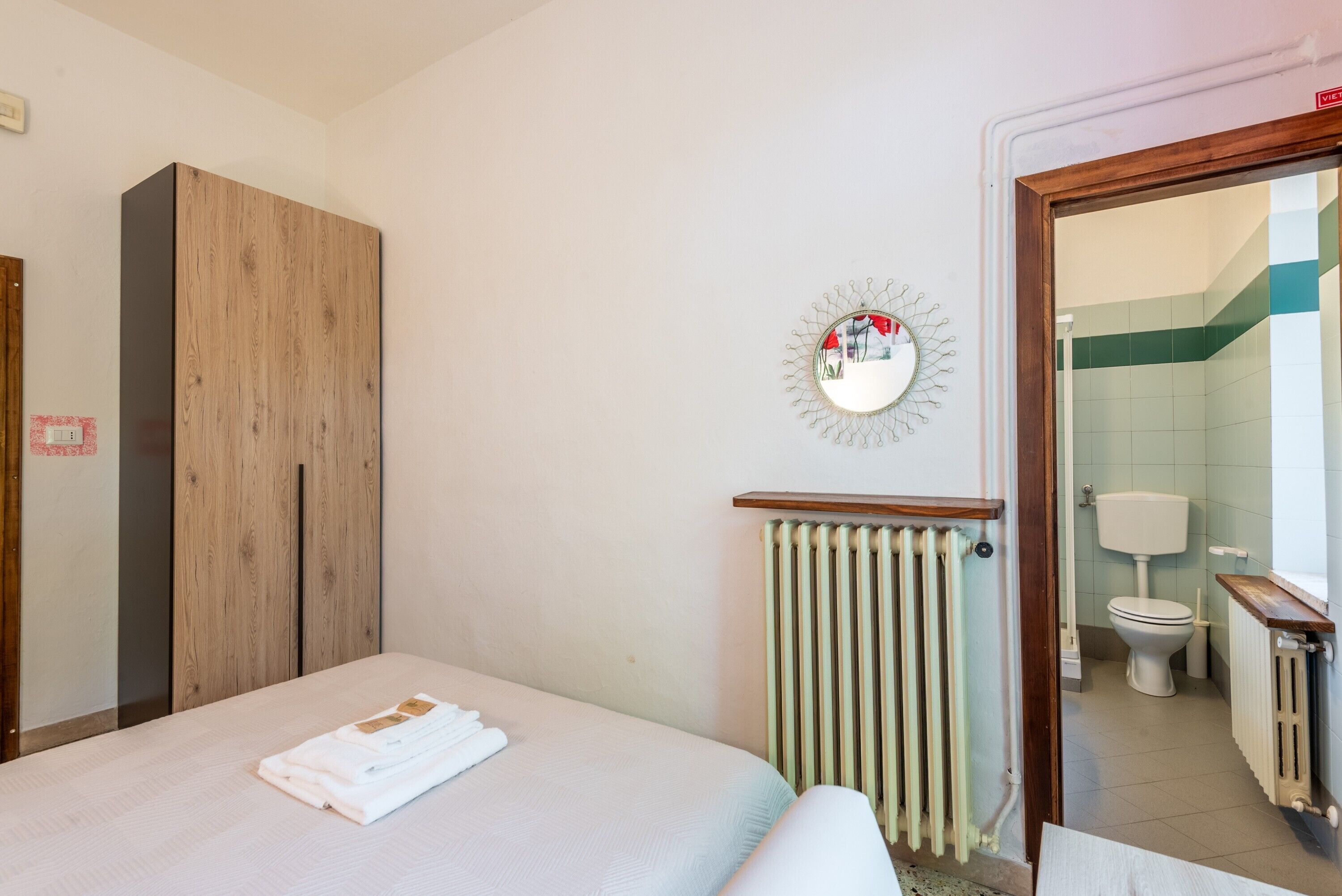 Quarto de hotel com cama, guarda-roupa de madeira, radiador, espelho e vista para o banheiro.