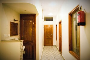 A narrow hallway with wooden doors, a tiled floor, a fire extinguisher mounted on the wall, and a window with a wooden frame. - Serenitysteay (Varanasi)