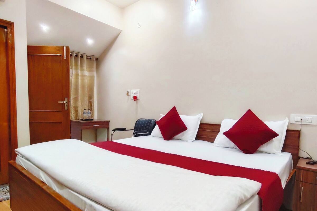 A neatly made bed with a wooden frame, a red and white bedspread, and two red pillows. There is a wooden nightstand with a lamp, a power outlet, and a wall socket. A door with a curtain is visible in the background.