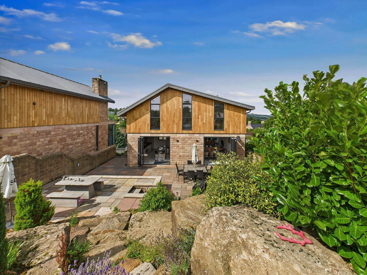 A modern house with a patio, stone pathway, and a garden with large rocks and shrubs.