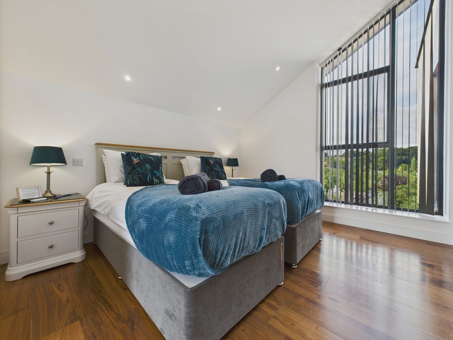 A bedroom with a large window, a bed with blue bedding, a nightstand with a lamp, and a wooden floor.