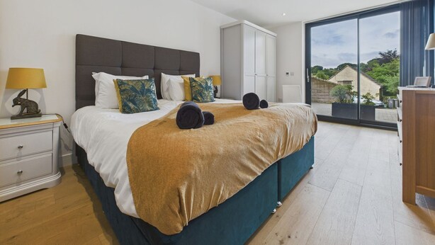 A modern bedroom with a large bed, a nightstand with a lamp, a dresser, and a view of the outside. - Oakwood House (Matlock)